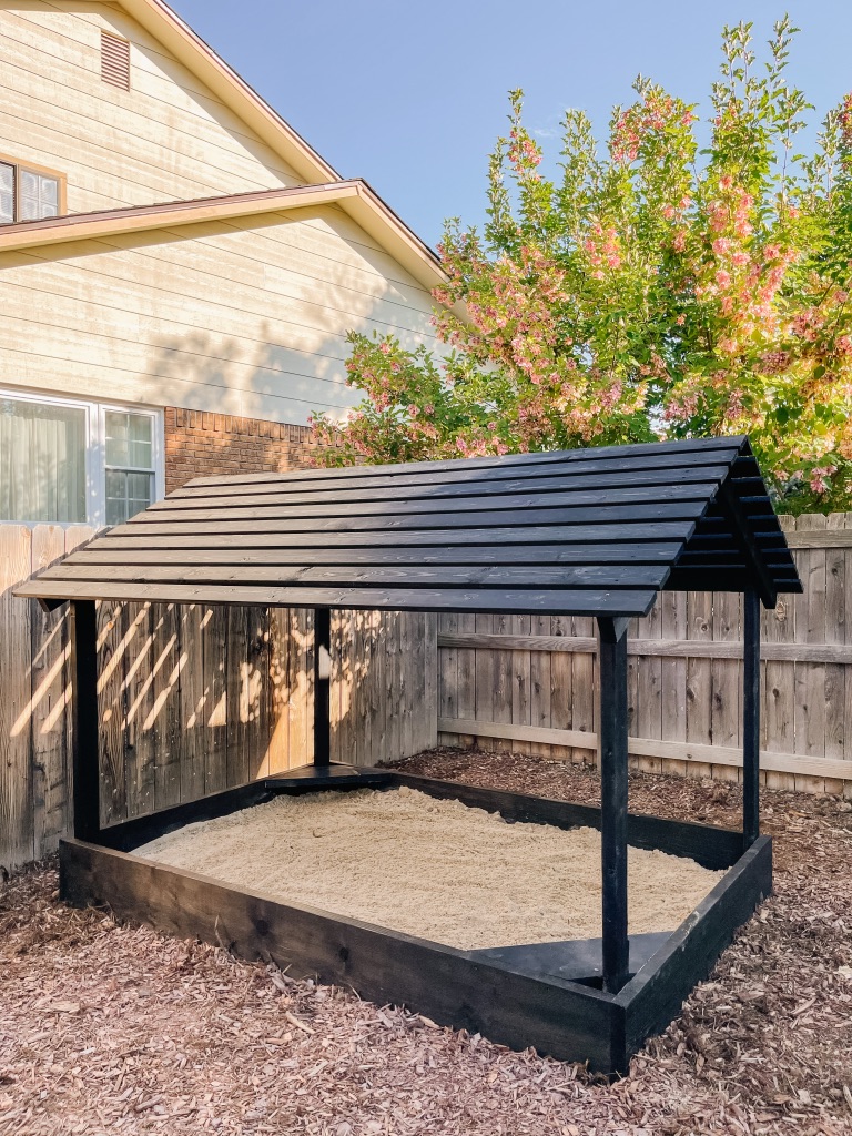 DIY sandbox with slatted roof and black wood stain in backyard