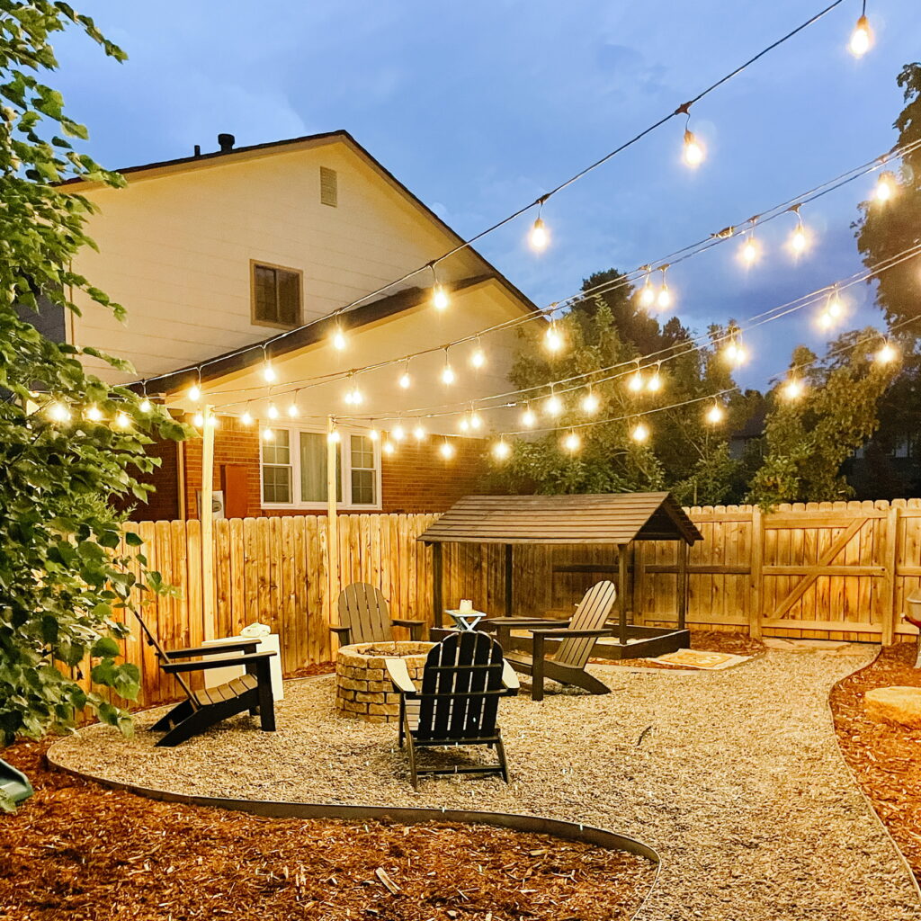Small backyard makeover with DIY Propane Fire Pit Area with Polywood Black Adirondack Chairs from Wayfair and DIY Sandbox with Slatted Roof at night with String Lights from Costco